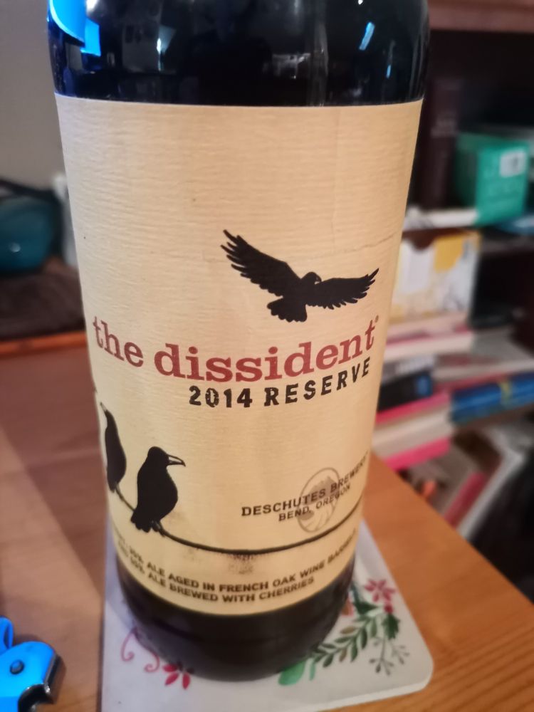 A 650ml bottle of The Dissident, a Belgian Bruno from Deschutes Brewing. The bottle is from 2014. The label shows three crows sitting on a wire though we only see two in this picture. A fourth crow flies away.  The words The Dissident 2014 Reserve are between the crows. 