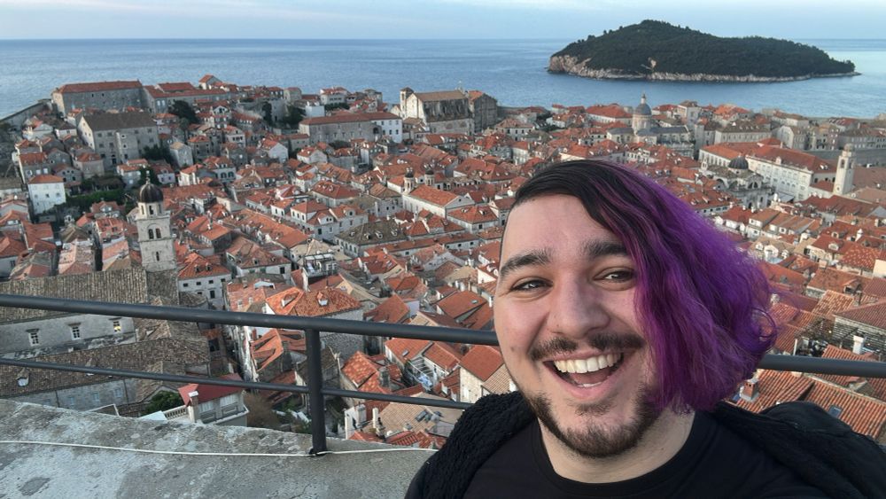 Ian is smiling while standing on the city walls above the old city in Dubrovnik Croatia at sunset. The city’s red tile roofs contrast with deep Ian’s deep purple hair