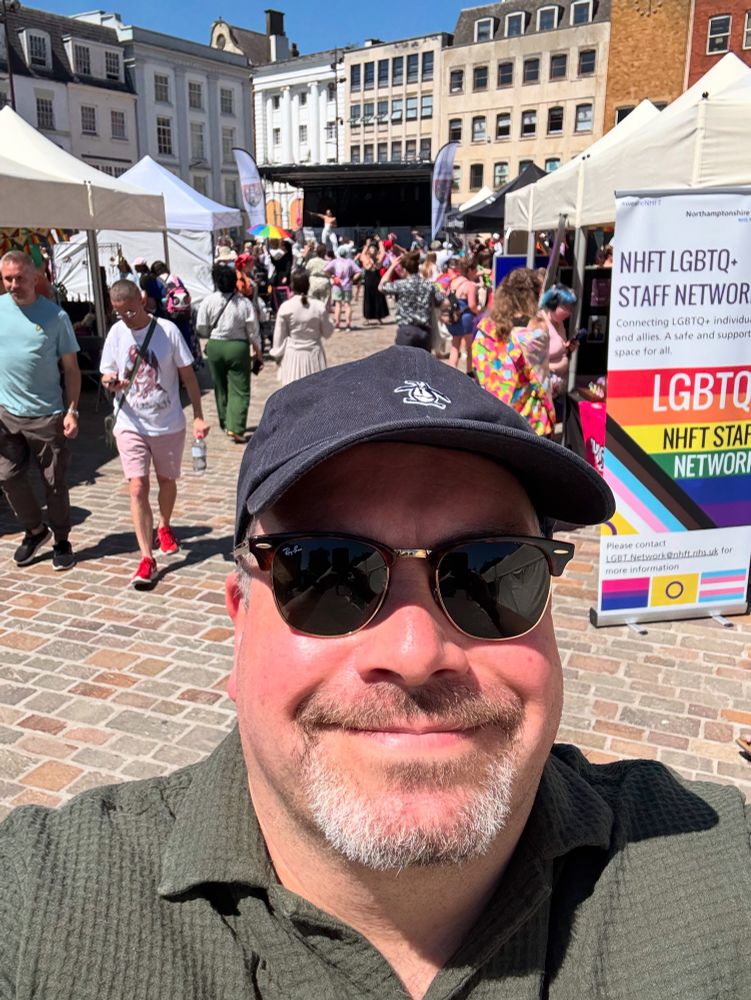 Selfie of a middle-aged man wearing a cap and sunglasses, in front of some pride-related stall and a performance stage in the distance