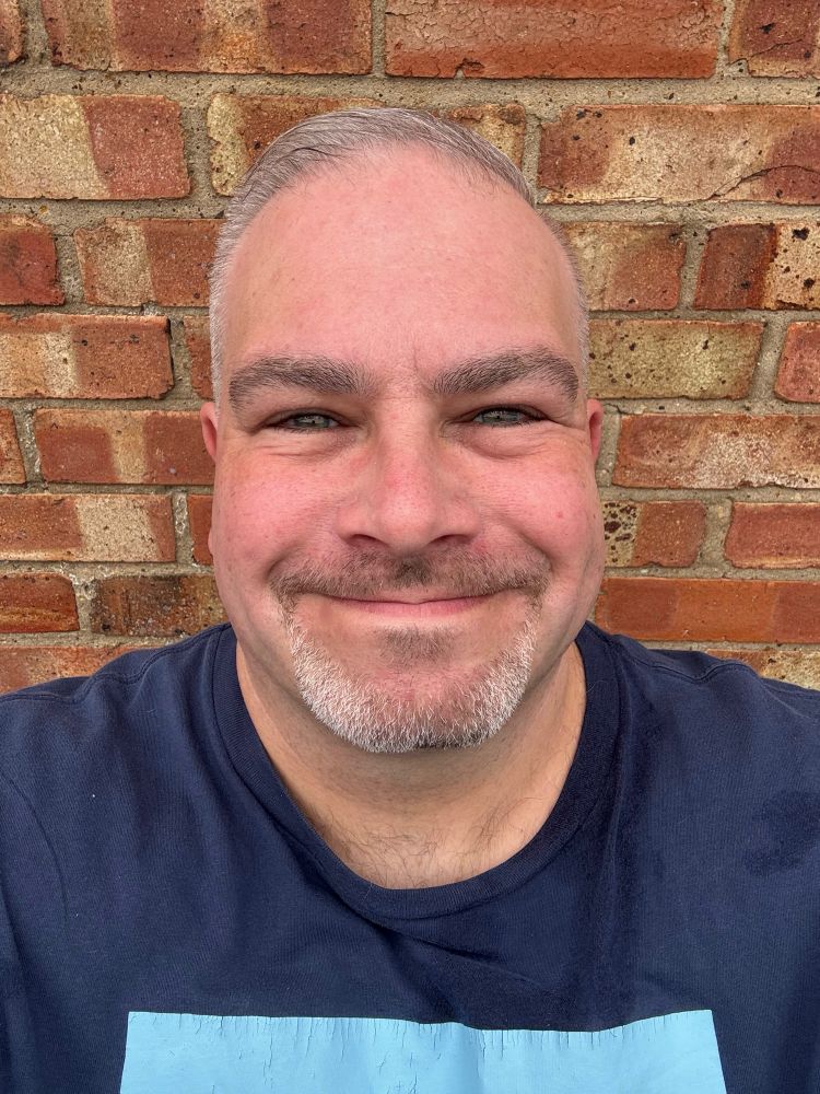 Selfie of a middle aged white man, wearing a navy t-shirt and standing in front of a brick wall