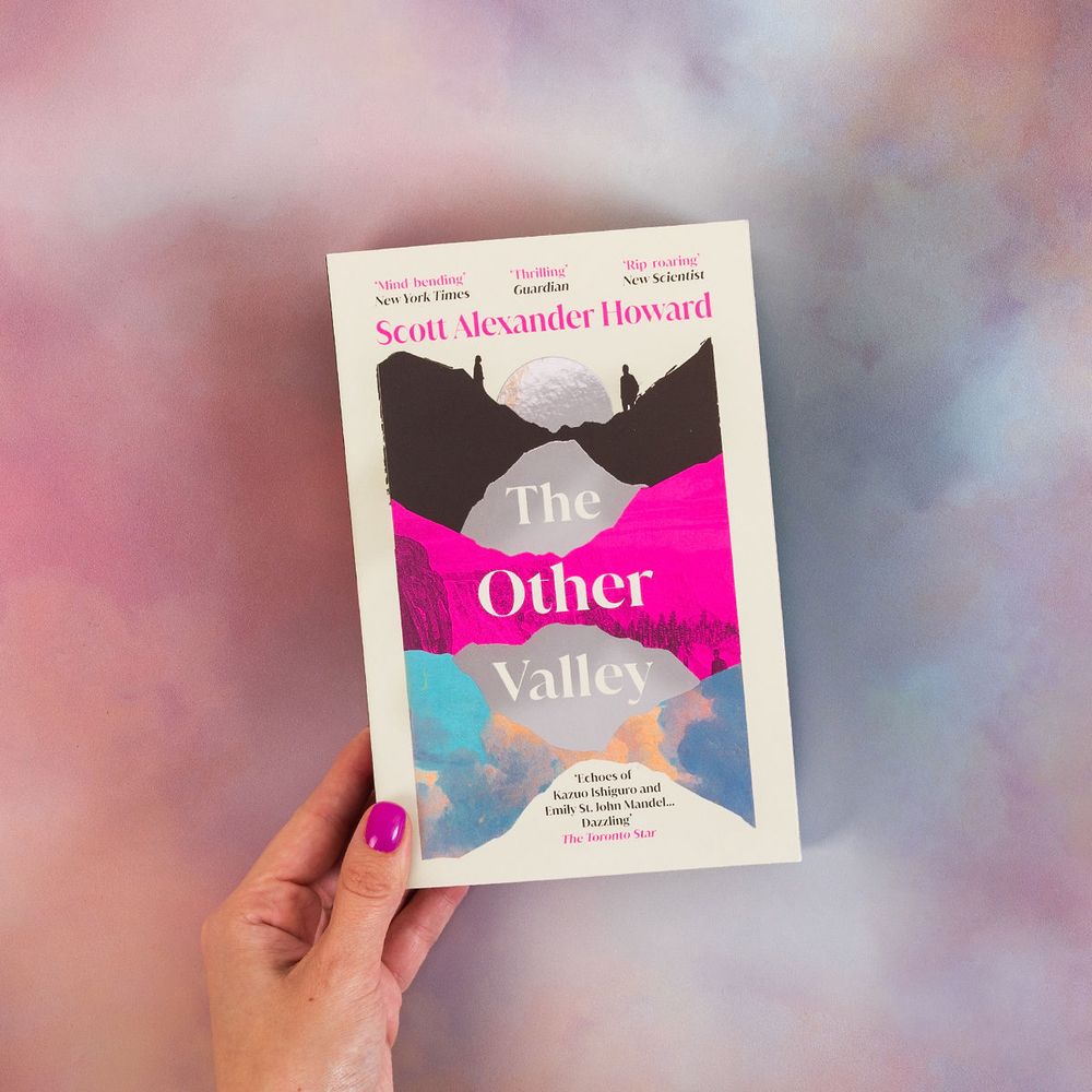 A photography of The Other Valley paperback (all neon pink, silver foil, with two figures in shadow standing either side of a moon) against a cloudy pink, blue and purple backdrop