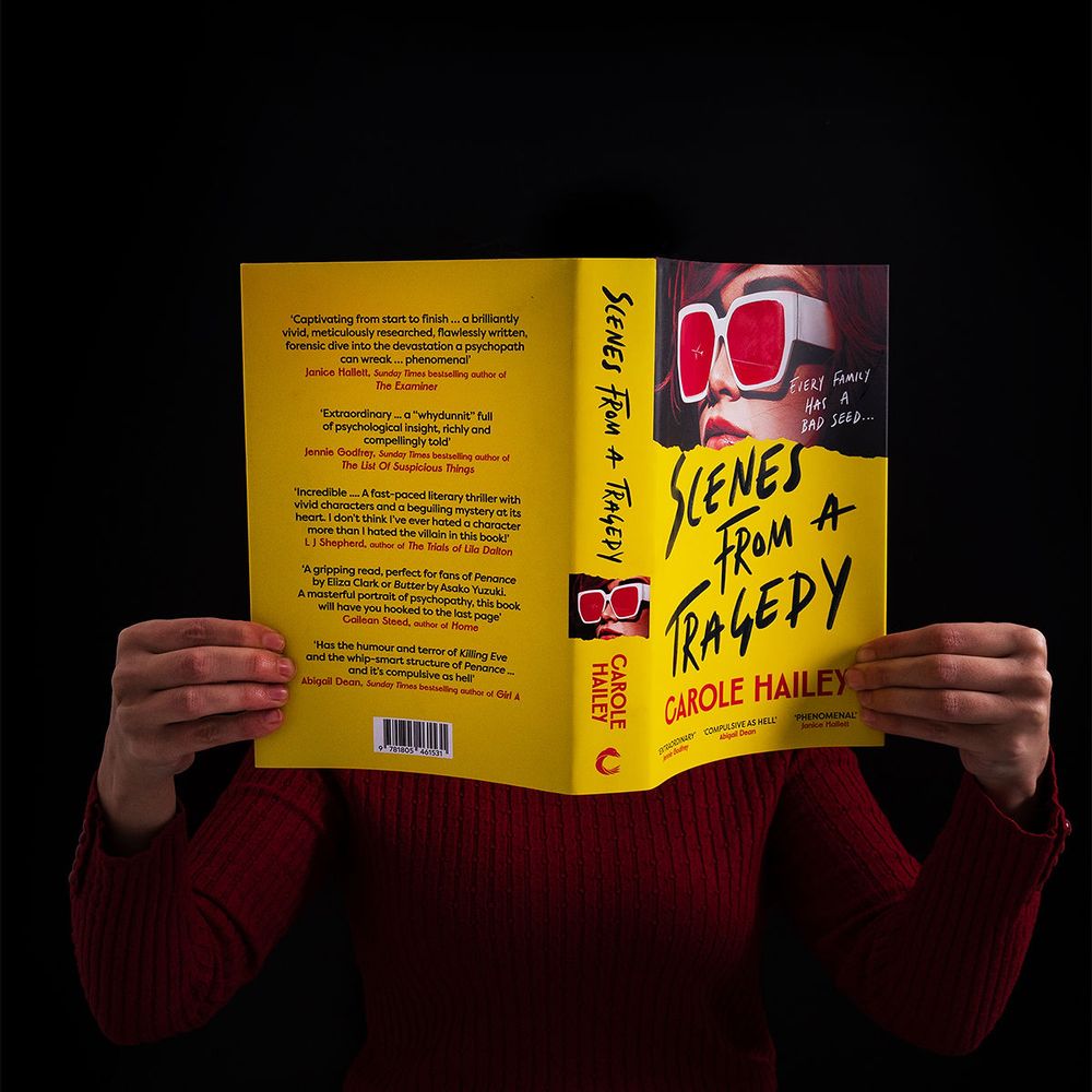 A bright yellow copy of Scenes From a Tragedy by Carole Hailey, held in front of a person's face, who is wearing a dark orange sweater