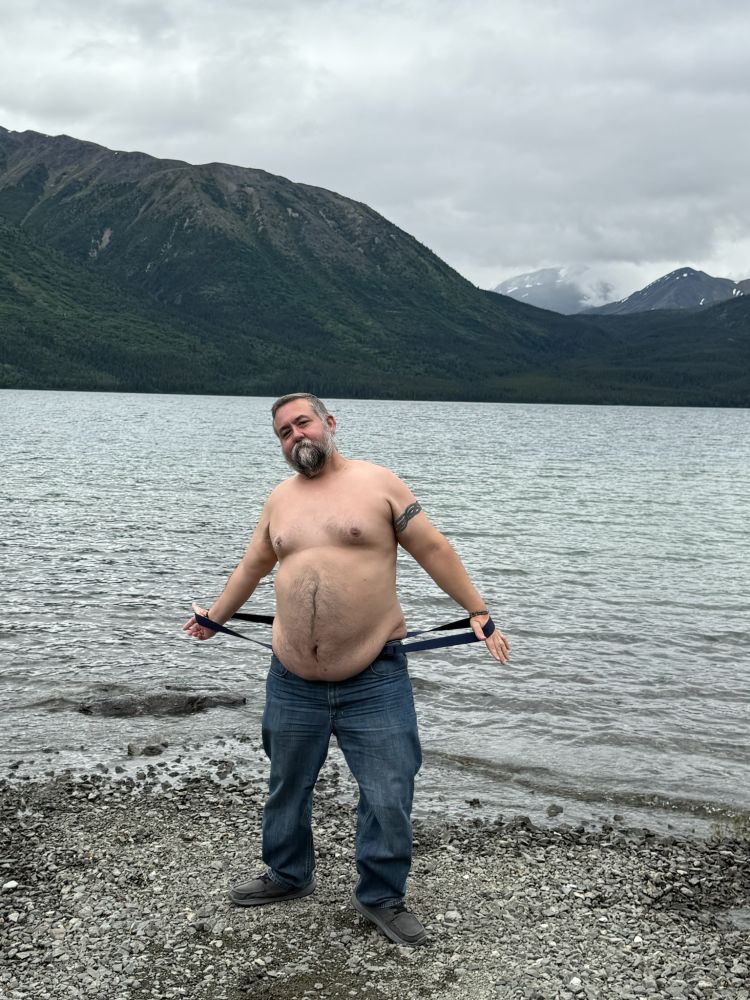 Chubby middle aged white guy with no shirt on in front of a lake in British Columbia, Canada.