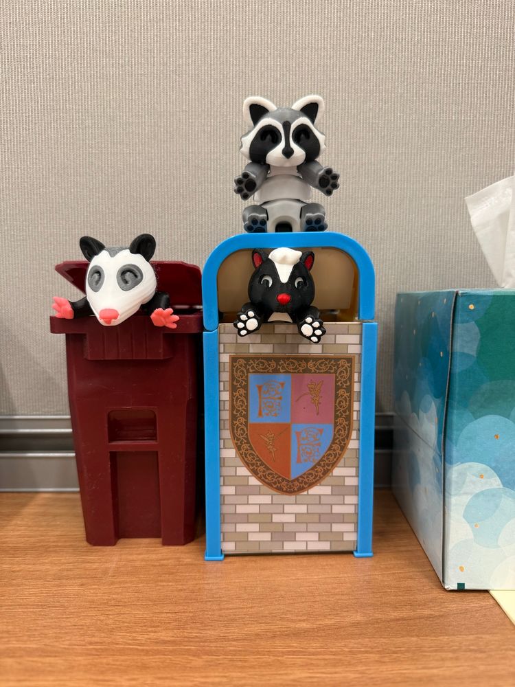 A possum, skunk, and raccoon 3D printed figures chilling on and in novelty trash bins on a desk.