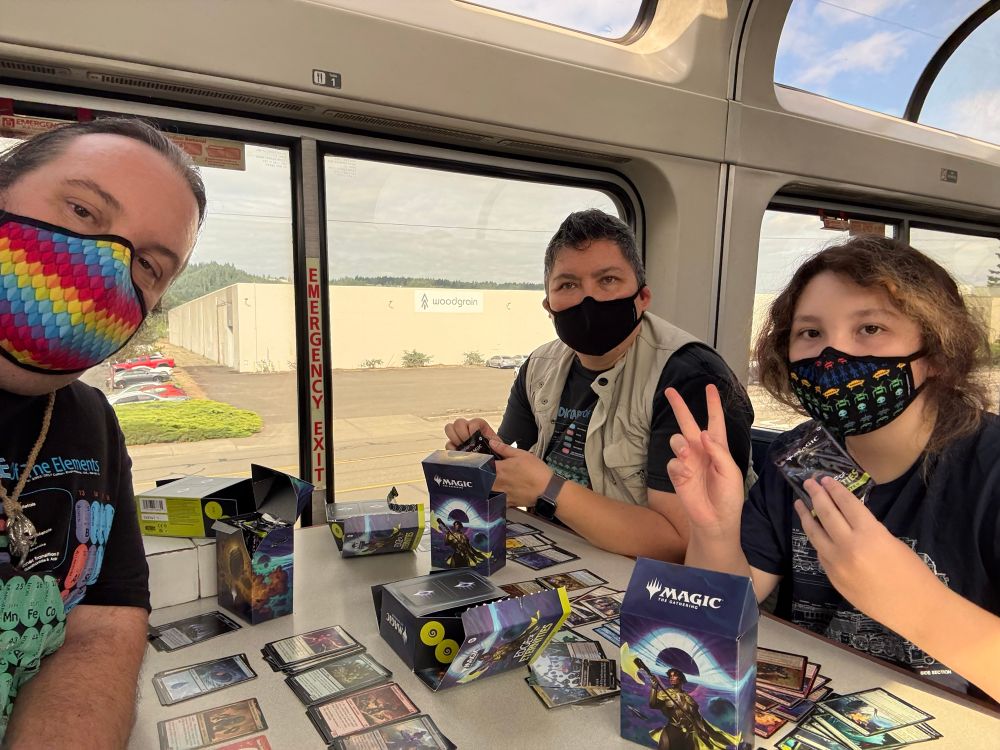 Three masked people sitting around a table in the observation car of a train, with Magic: The Gathering cards spread out on the table, and boxes of the Edge of Eternities set in the center of the table.