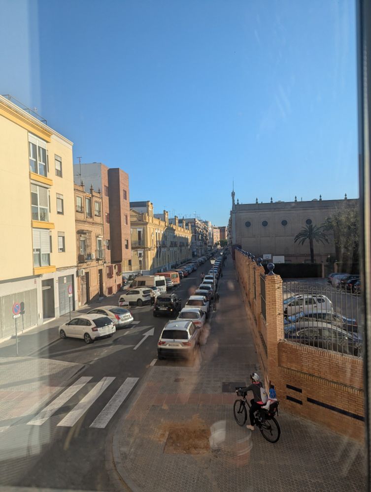A view from the hostel in Sevilla, a quiet side street, some parked cars, and a cyclist, under a big blue sky.
