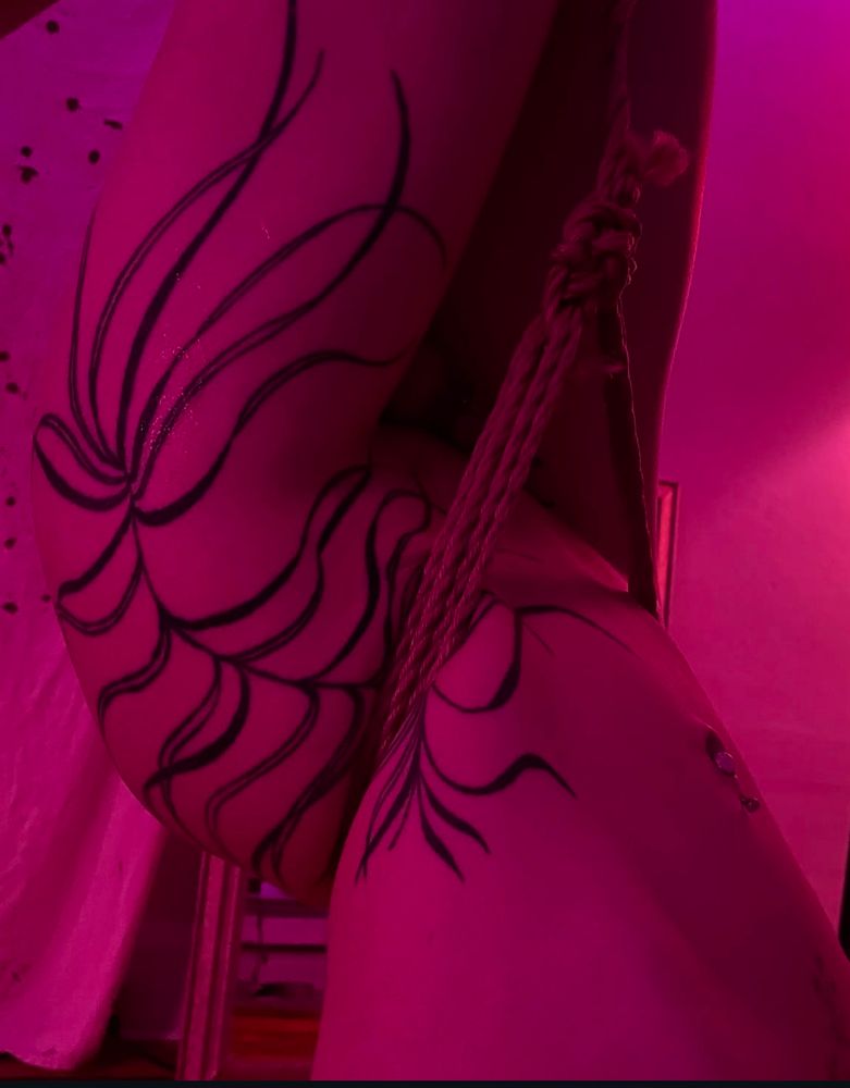 in a slutty pink overtone we see a tattooed femme upside down with rope cinching her waist as she hangs. The tattoo featured is a large black and grey botanical palm frond or fern like design hand drawn on and tattooed by @jerktattoo it stretches from mid waist height onto the hip, butt and thigh. 