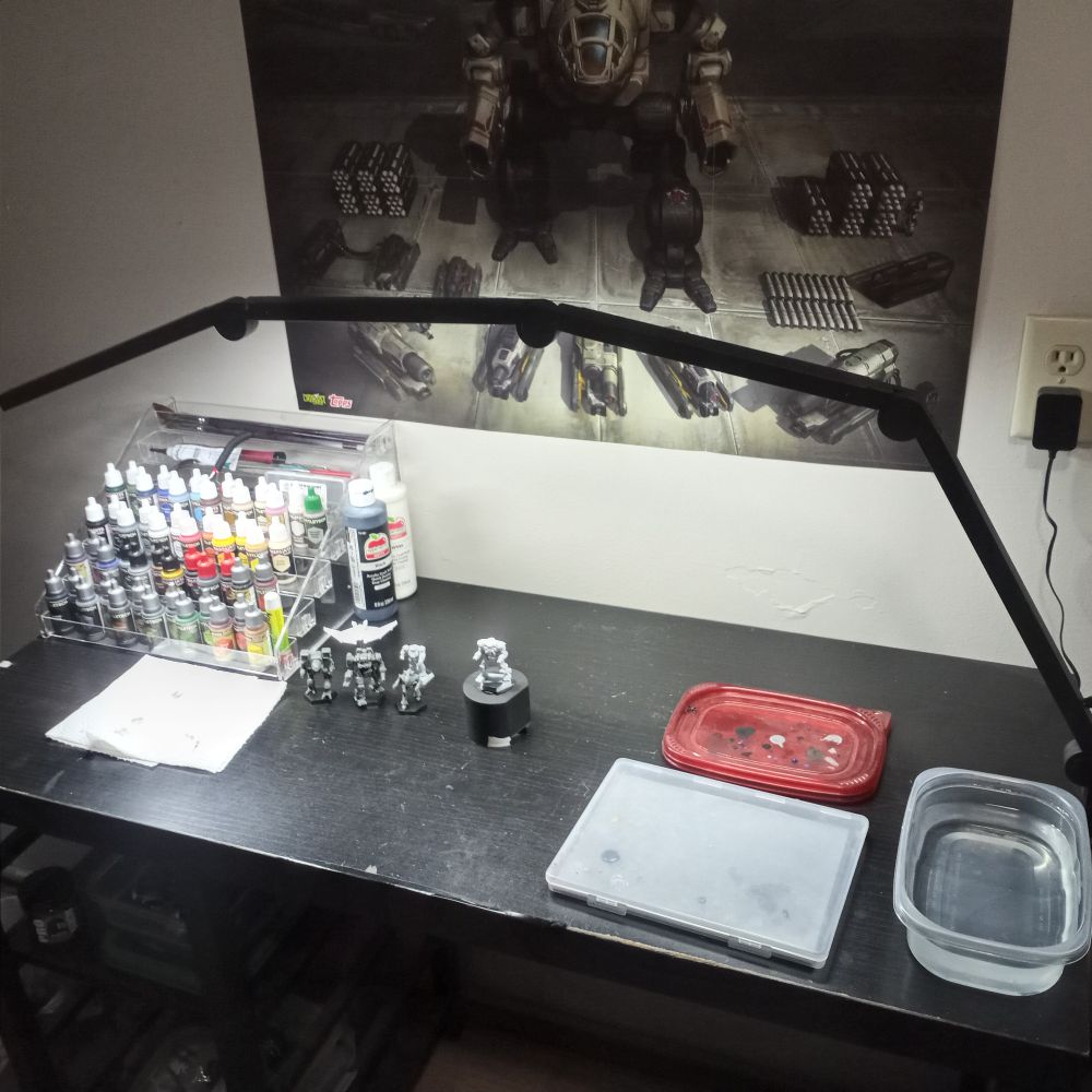 A black painting table with an array of paints, some miniatures, water, paper towels, etc. A poster of a Timber Wolf is in the background.