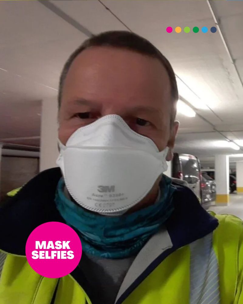 Ein Mann mit kurzen, hellen Haaren steht in einer Tiefgarage. Er trägt eine reflektierende gelbe Sicherheitsweste, ein blaues Halstuch und eine 3M Aura N95-Maske. Der Hintergrund zeigt Parkplätze und Autos. Ein pinker Kreis mit weißer Schrift unten links enthält den Text „MASK SELFIES“. Oben sind sechs bunte Punkte und das „whn.global“-Logo zu sehen.