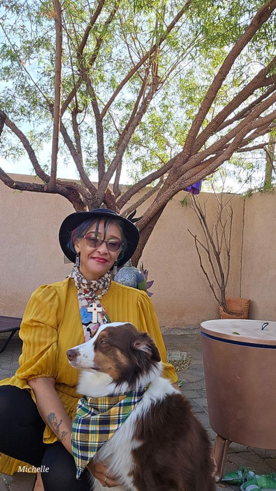 I am in a mustard colored dress with a black hat on, crouched behind my red tri-color Australian Shepherd, who is wearing a matching plaid handkerchief around his neck. We are posing in front of a tree and a wall. 