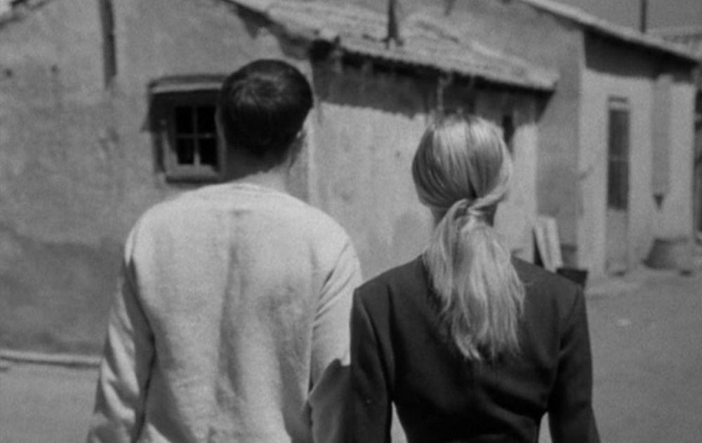 A black-and-white image from the film La Pointe Courte, depicting a man and a woman walking side by side through a rustic village scene. The man is wearing a light-colored shirt, and the woman has her blonde hair tied in a ponytail, wearing a dark jacket. Their backs are turned to the camera, and the setting includes weathered buildings with a textured, aged look.