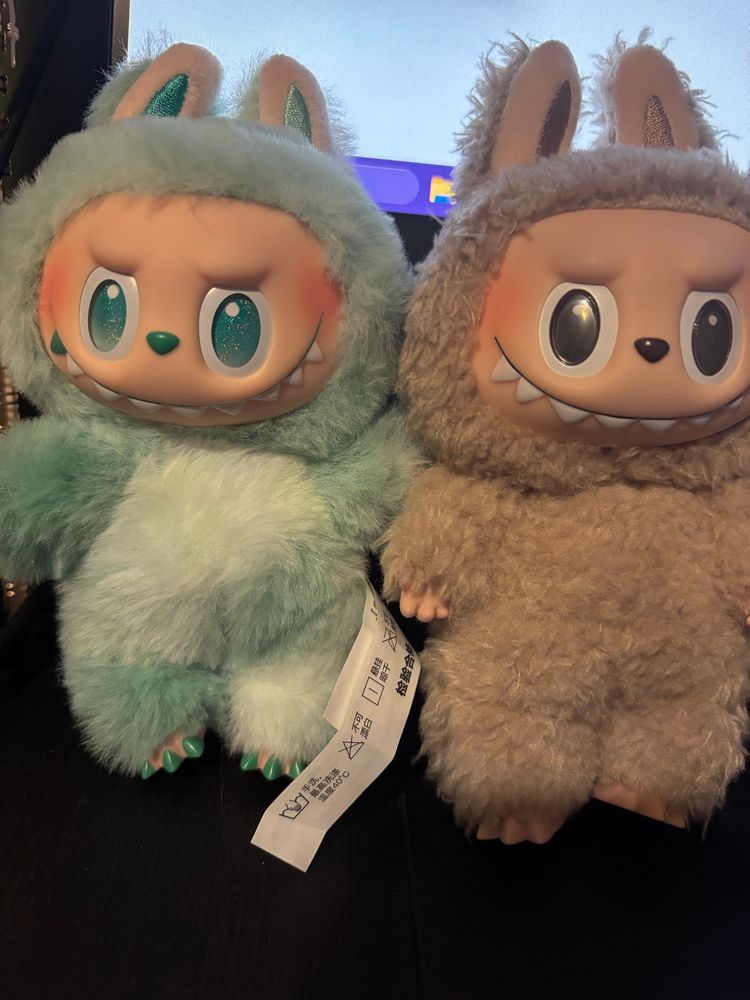 Two labubus standing next to each other. One is green with green eyes and the other is a toffee color with brown eyes.