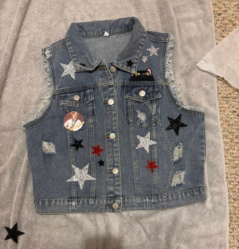 A denim jacket with various silver, black, and red star patches, a cat patch, a silver chain, and a button with a picture of a man playing a guitar 