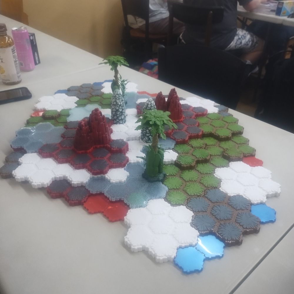 A Heroscape terrain map featuring grey rock, blue water, white snow, green grass, translucent ice, and red lava hex tiles. There are jungle and evergreen trees with red rock outcroppings in the center