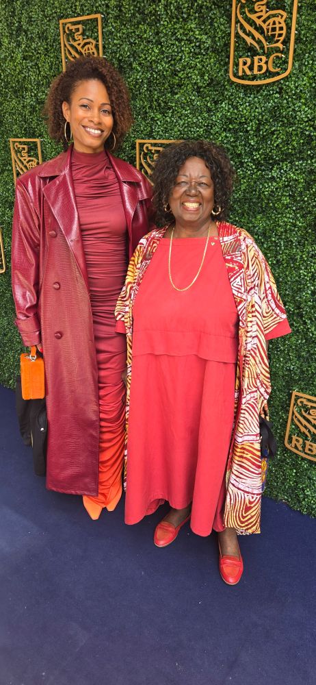 2 Black women smiling - one in an ombre burgundy to orange dress and burgundy trench coat, the other in a red-orange dress with a white/yellow/red jacket 