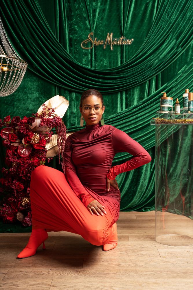 Bee in a red to orange ombre long sleeved dress, in a squat pose in front of a green SheaMoisture backdrop