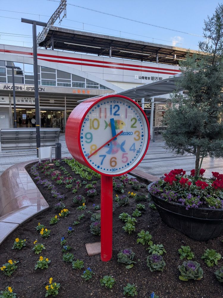 駅前のロータリーにある花壇とそこに突き刺さるように建てられた時計。
花壇には等間隔に黄色や青、紫の花が並んでいる。
時計は赤色で、文字盤は青や黄色などカラフル。真ん中に二匹の魚のイラストが描かれている。
