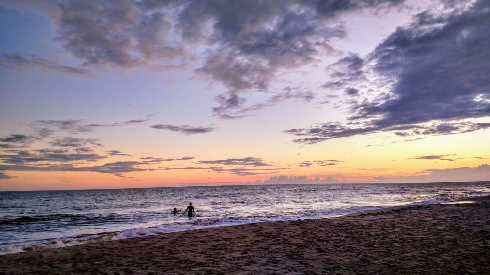 Sunset on the Atlantic Ocean, the silhouette of two people playing in the water stands against the calm rolling waves, an orangey-pink glow on the horizon, and light grey clouds pass by above.