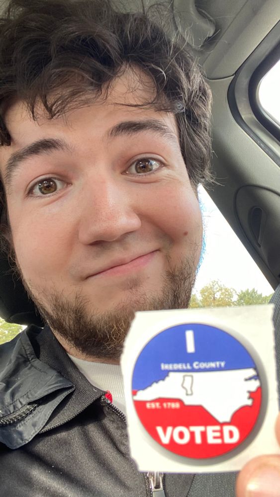 A selfie of I, Michael Henson, holding up an “I voted” sticker for my local municipal election. I have dark brown hair with hazel eyes, a shortly trimmed beard to age my eternally youthful face, and am in dire need of a haircut.