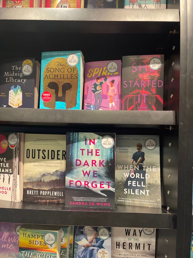 Photo of a bookshelf at Vancouver International Airport with my novel IN THE DARK WE FORGET among others such as SONG OF ACHILLES, FIVE LITTLE INDIANS, and THE MIDNIGHT LIBRARY, all with stickers that read “2 for $40”