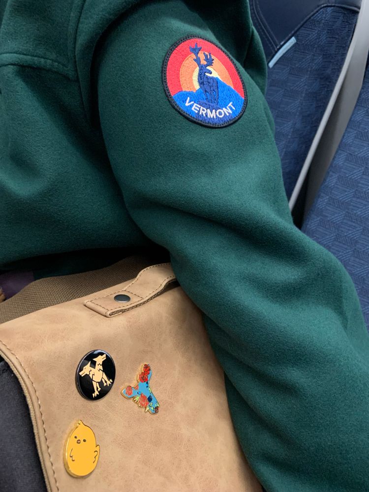 Image shows green coat with a Vermont patch on the arm. The bag is light brown with 3 pin badges on it. 