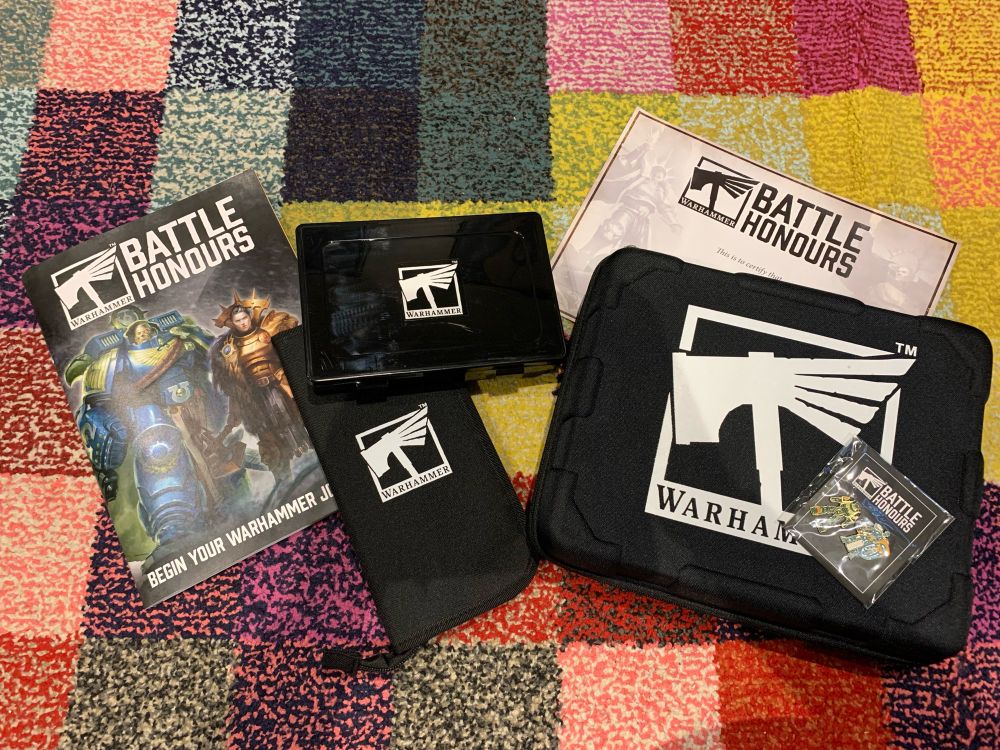 Image shows 6 items on a checkered rug. 
It is the Battle Honours that Warhammer stores have to encourage new players to the hobby. It has a range of tasks from collecting, building, painting and more. If you complete the tasks you get rewards.

Shown are:
- The magazine to keep track of tasks
- Box for your loose bits
- Paint brush pouch
- Pin badges
- Storage box (after completing 30 tasks)
- Certificate 
