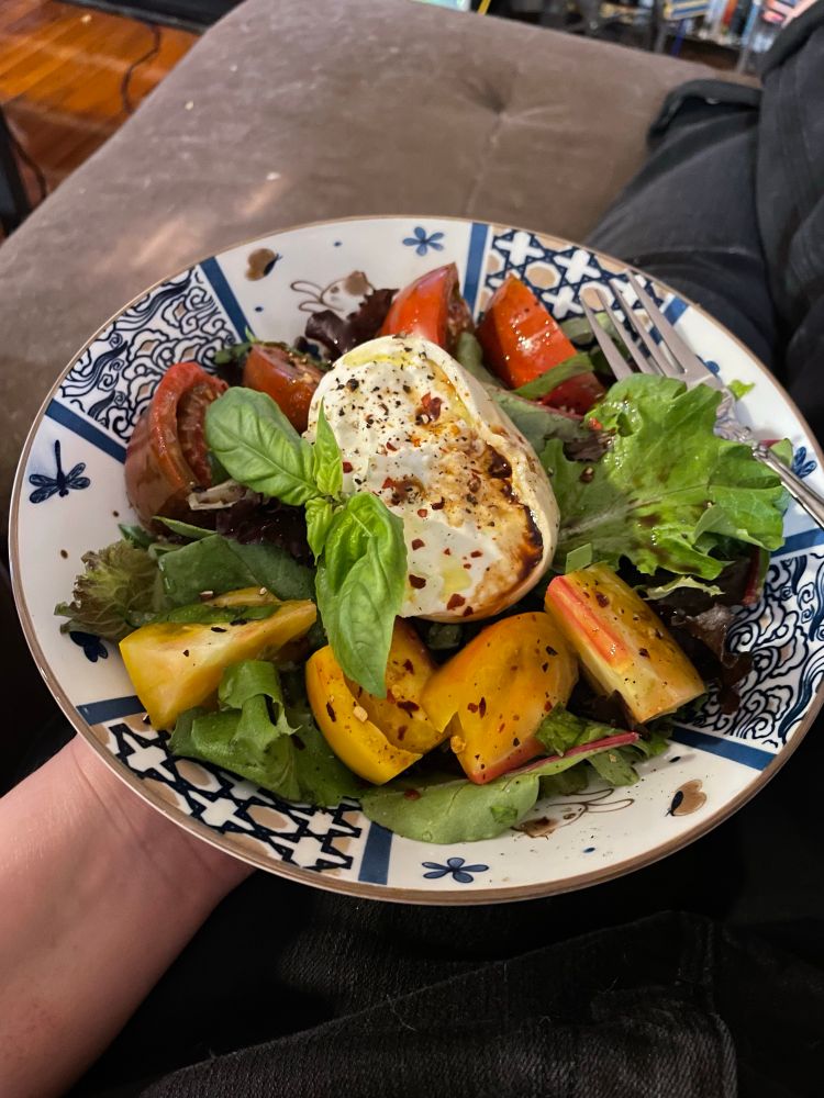 A plate with salad greens, two kinds of tomatoes (one yellow and one red) cut into wedges, a half of a ball of burrata, some fresh basil, some fresh cracked black pepper, a sprinkling of red pepper flakes, a splash of balsamic vinegar, and a light drizzle of olive oil.