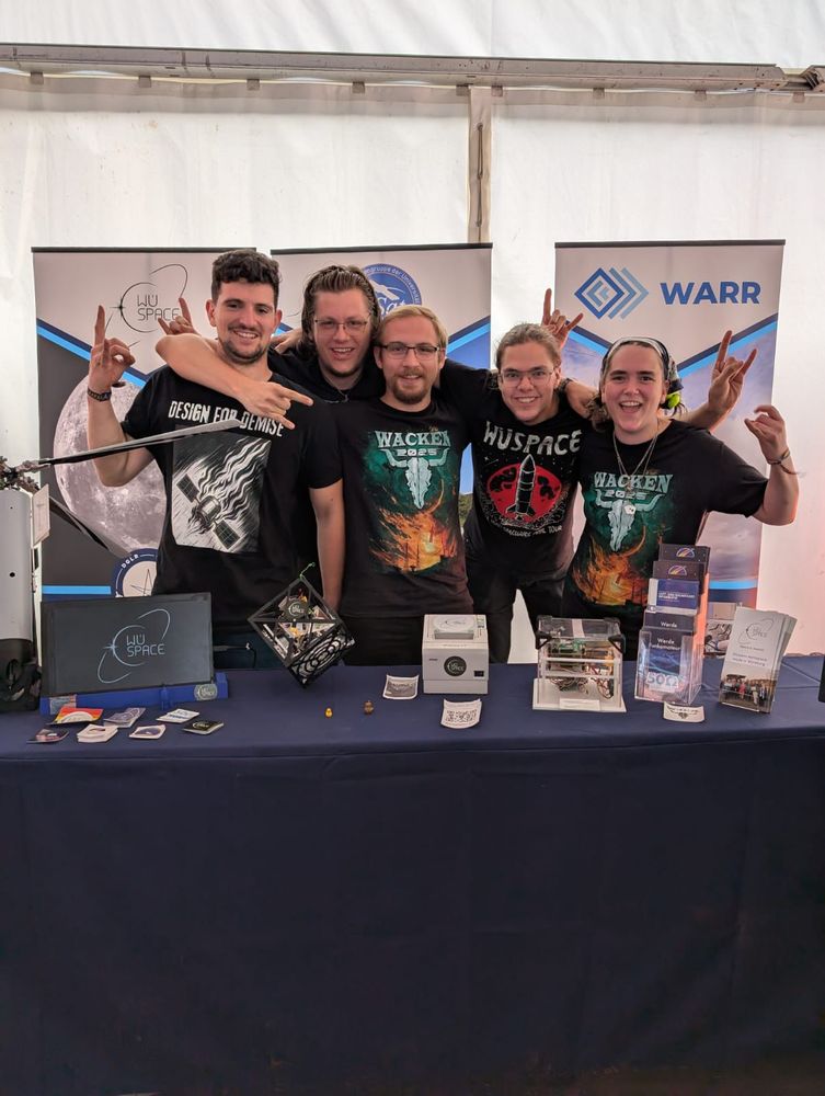 A group photo of 5 members of WüSpace behind their exhibition table, which houses different aerospace hardware from various projects of WüSpace.
