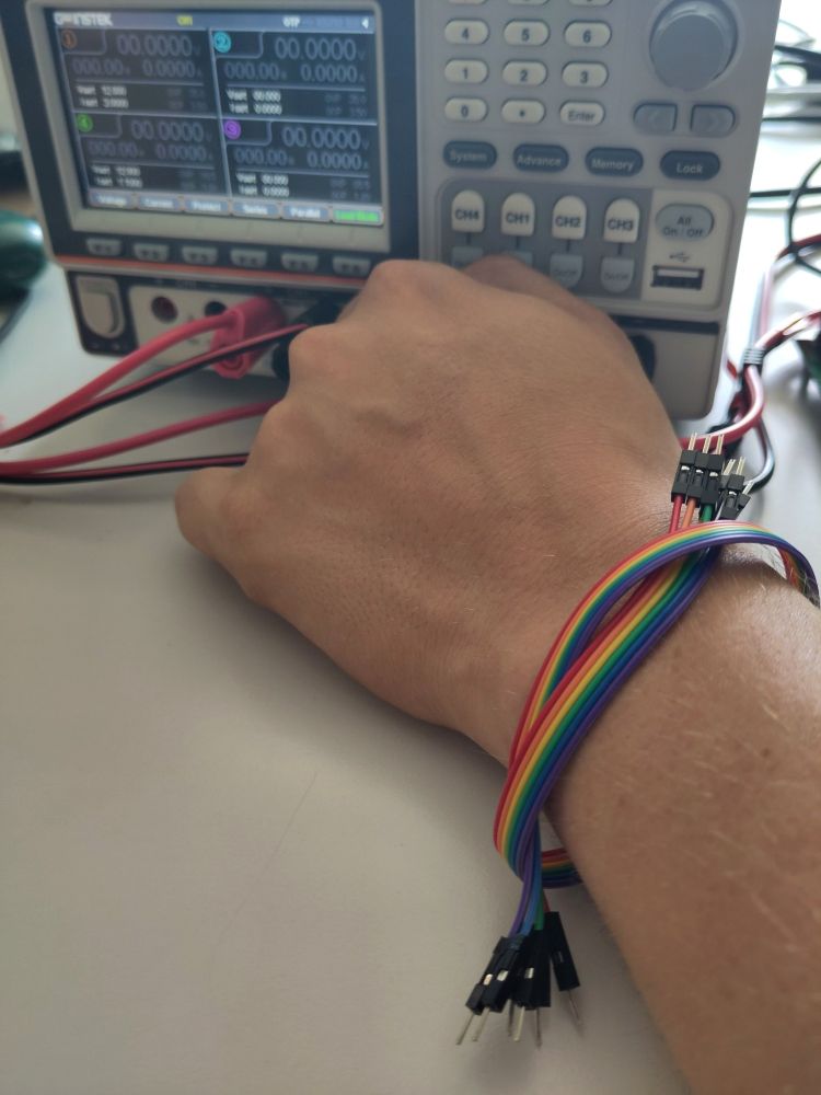 Picture of the left hand of one of our members in front of a lab power supply. They are wearing a bunch of dupont wires in rainbow order around the arm as a bracelet.
