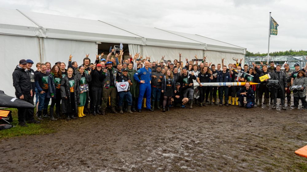 A group photo of approximately 60 people in front of a big pavilion. In the middle is ESA astronaut Alexander Gerst. Some people are holding small rovers, a small rocket, or laptops. Many people wear merchandise of Wacken Open Air, and are gesturing the Metal Sign.

