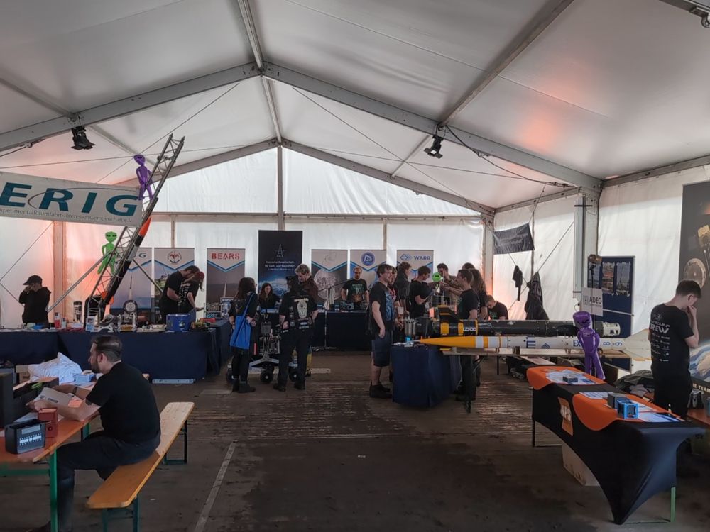 Wide angle photo of the inside of a big pavilion, giving an overview of the different exhibits in the Space Camp. You can see Small Rockets and Rovers in the back, with some benches and tables in the front. A couple of visitors and a couple of exhibitors are scattered around.
