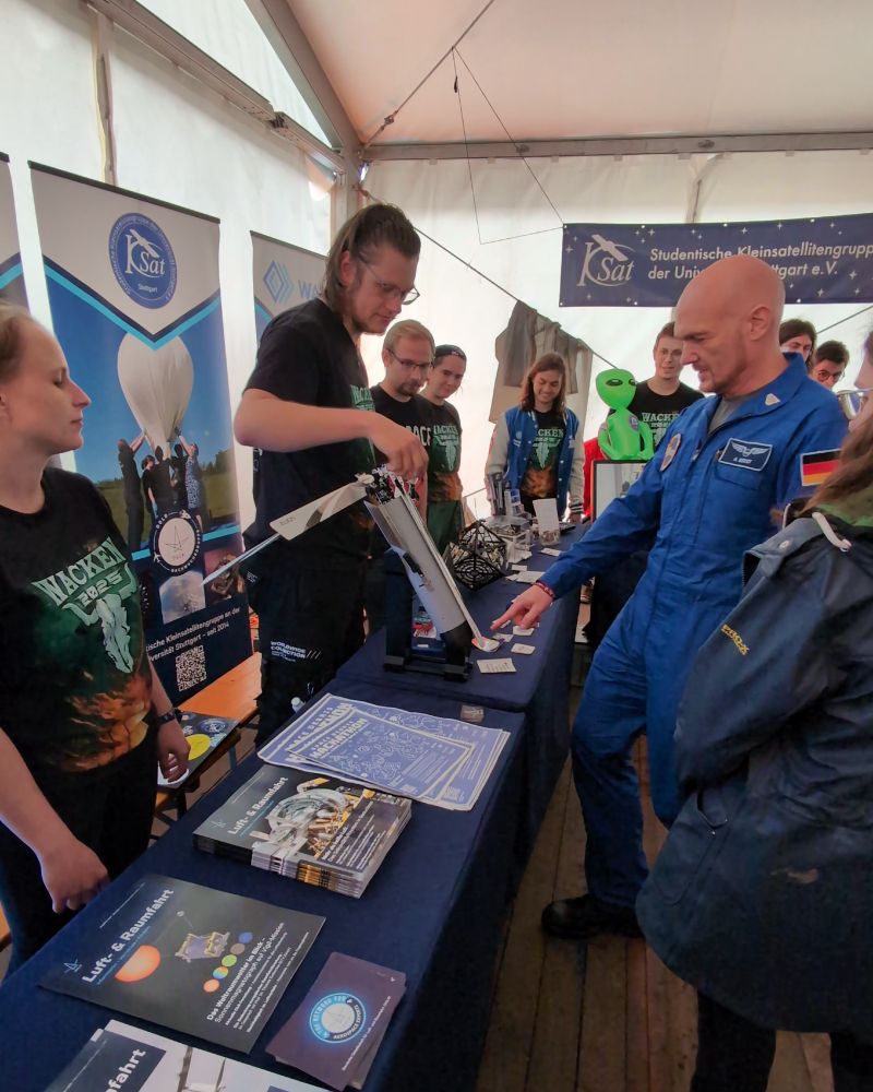 Two exhibition tables covered in aerospace hardware and informational material from DGLR and WüSpace. A member of WüSpace on the left explains an experiment to Alexander Gerst, who is stood on the right.
