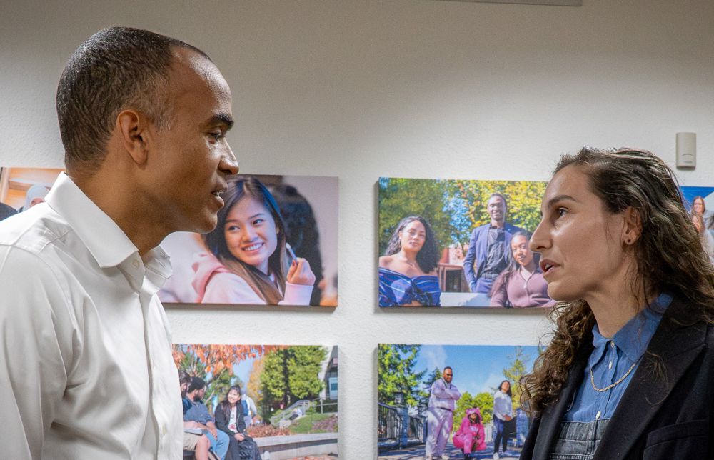A man in a white shirt (left) is facing a woman with long, wavy hair (right) who is wearing a dark jacket over a blue shirt, and they are engaged in conversation. They are indoors, and the wall behind them is decorated with multiple large photographs of people in various outdoor settings.