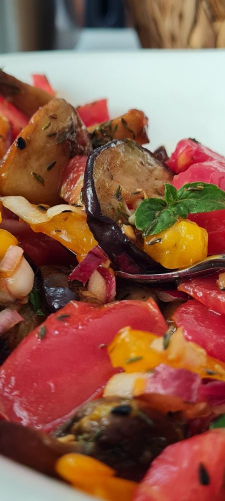 Ein Löffel steckt in einem bunten Salat aus roten und gelben Fleischtomaten, Melanzani und Champignons, mit Kräutern. Ein Basilikumkrönchen ist rechts in der Mitte des Bildes. Der Salat wurde in weißer Keramik angerichtet.