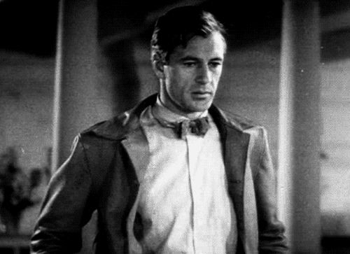 Gary Cooper in a 1930s movie wearing a brown leather or suede jacket, a white dress shirt and a bow tie, blinking and then looking downward bashfully.
