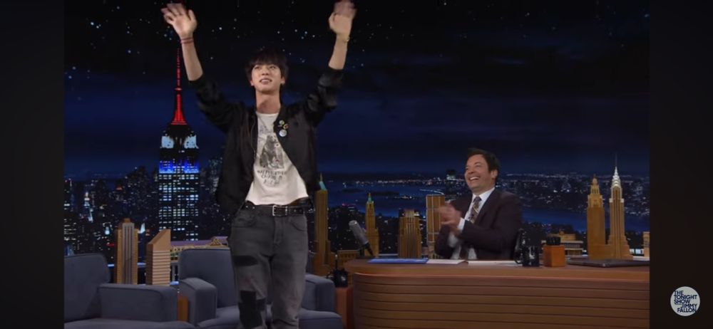Jin of BTS waving at the audience before taking a seat to begin his interview with Jimmy Fallon, who’s sitting behind his hosting desk.