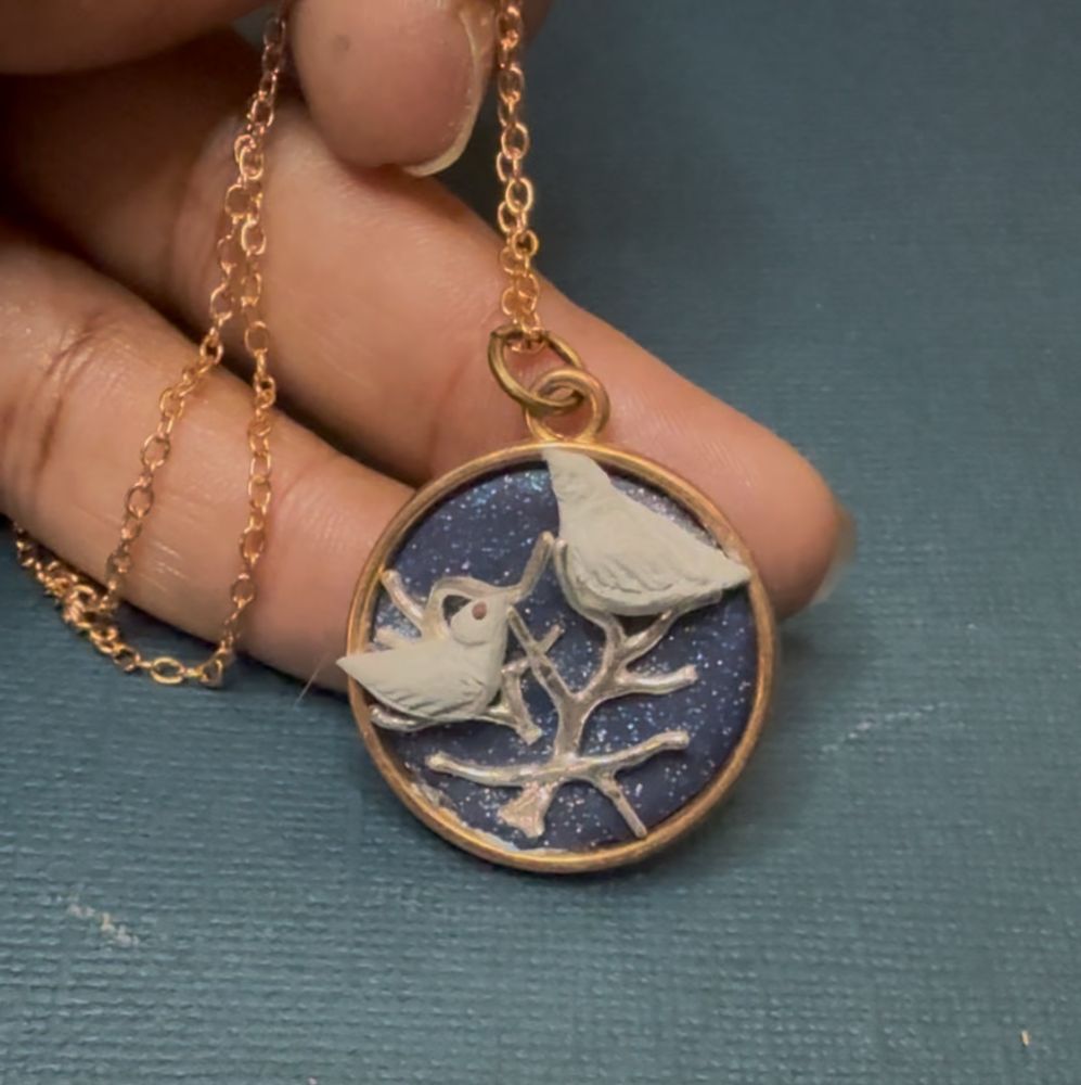 Necklace pendant made of sculpted clay, repurposed silver pieces and a gold bezel on gold chain. Two white birds perched in silver tree branches on a blue sparkling sky background
