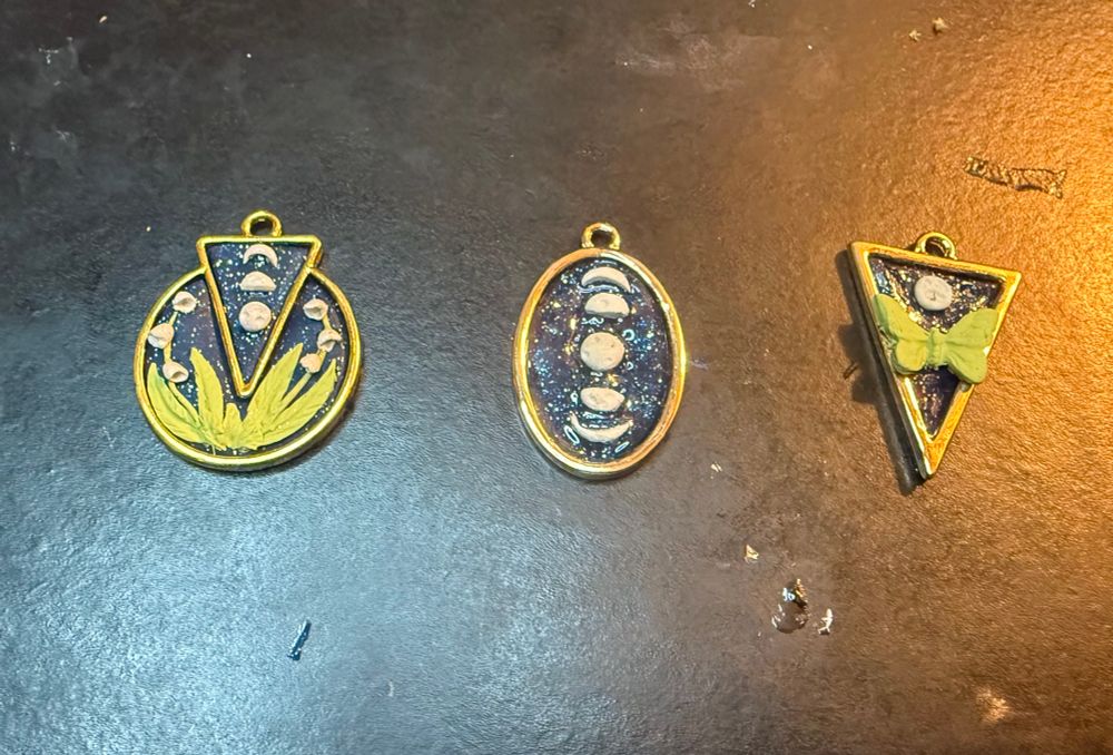 Three necklace pendants in gold bezels: one depicts three moon phases over a lily of the valley on a dark blue sparkle background, and other shows five moon phases on a dark blue background and the final has a green moth with a full moon on a dark blue background 