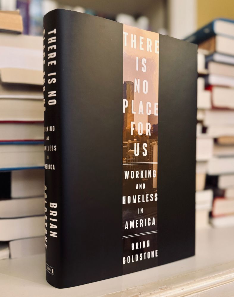 A hardcover copy of There Is No Place for Us: Working and Homeless in America by Brian Goldstone. The black cover features a vertical strip with the title and a photo of Atlanta’s skyline.