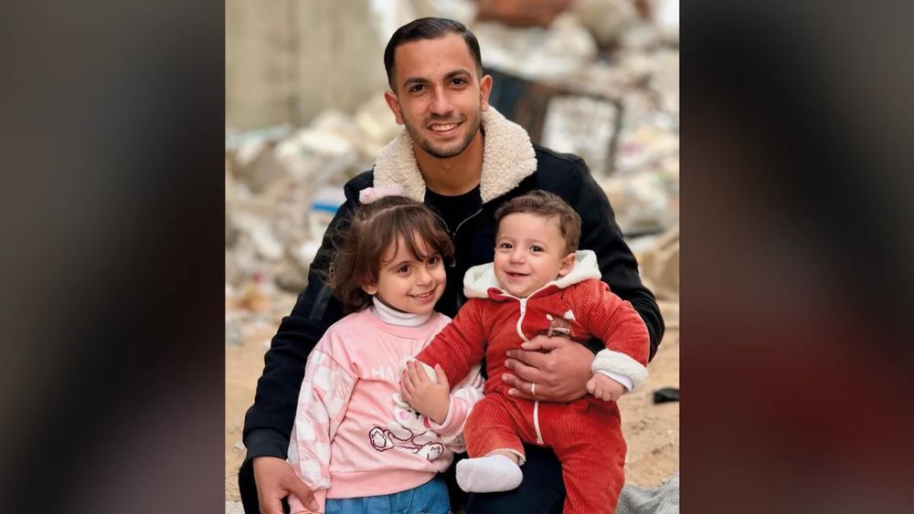 Anas al-Sharif, smiling, sits outdoors holding his young daughter Sham on his left and his infant son Salah on his right. Both children are smiling; rubble is visible in the background.