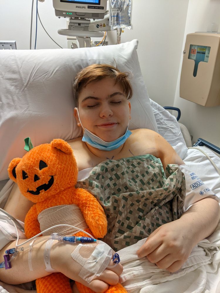 White person with brown hair laying in a hospital bed with bliss on their face. They have an IV in their arm and are holding a stuffed bear with their own bandages over its chest. 