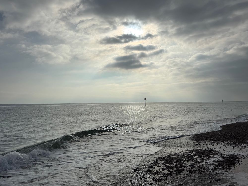 Silver-lit sea under a cloudy but bright autumn sky. A wave furls gently. A breakwater post on the horizon.