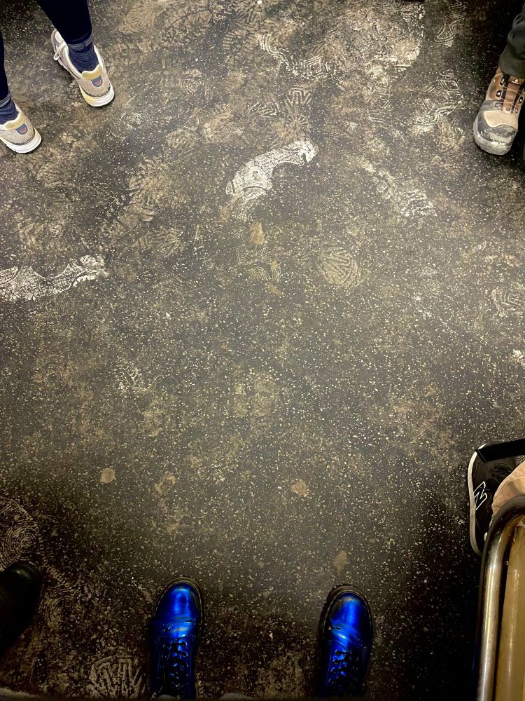 a photograph of the various layers of footprints left behind due to recent snow and salted streets or perhaps from construction and such, overlayed on the speckled flooring of the subway car, with a few other passengers feet cropped into the image, including a pair of shiny metallic blue dr. martens boots