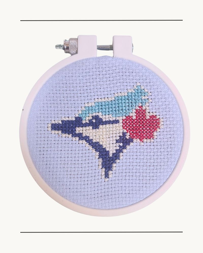 The logo of the Toronto Blue Jays cross stitched on a light blue fabric and white embroidery hoop