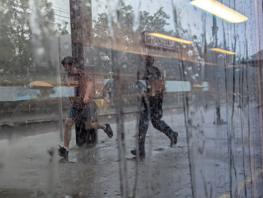 View out of the train window in Mechelen Nekkerspoel. Rain is streaming down the window while two people outside run to get on the train as quick as they can. Warm yellow light from the carriage is reflected and appears where the sun should be. 