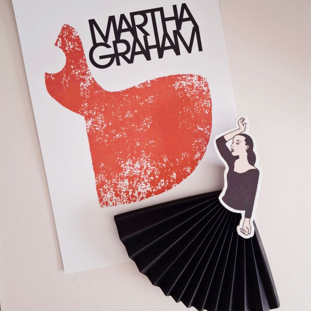 A black paper fan forms the dress of a Martha Graham figure. There is also a small art print in the background with an abstract depiction of her dancing.