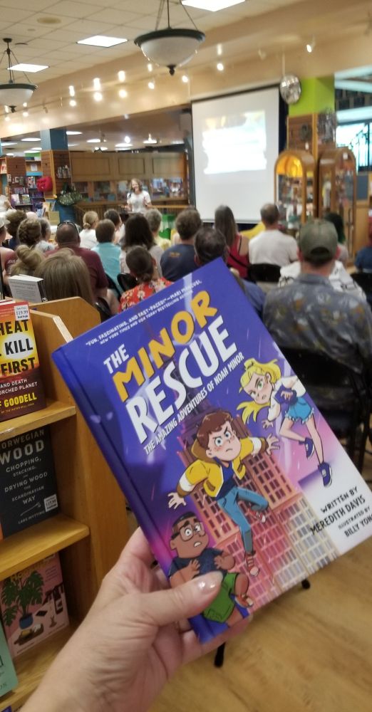 Copy of the book THE MINOR RESCUE written by Meredith Davis and illustrated by Billy Yong in front of a crowd of people at BookPeople in Austin TX sitting listening to Meredith Davis talk about the book. 