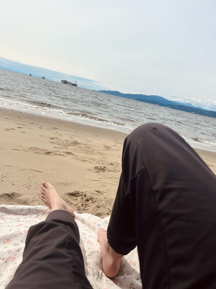 Light skinned person in black pants and no socks on a plant patterned picnic blanket on the beach facing the waves and mountains in the distance 
