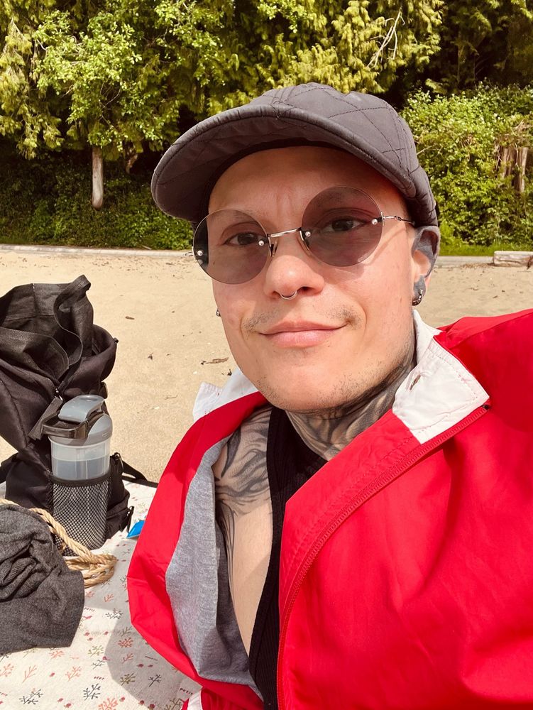 Light skinned person in a black cap, tinted glasses and red jacket leaning on a plant printed picnic blanket on the beach, trees behind them. Beside them sits a black backpack with a water bottle, sweater and rope.