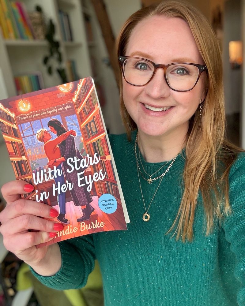 A woman holds a book with two women embracing on the cover. WITH STARS IN HER EYES by Andie Burke